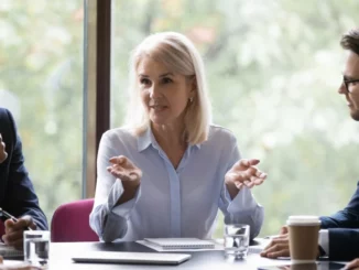 Frau sitzt mit anderen Menschen am Kopf eines Tisches in einem Meeting